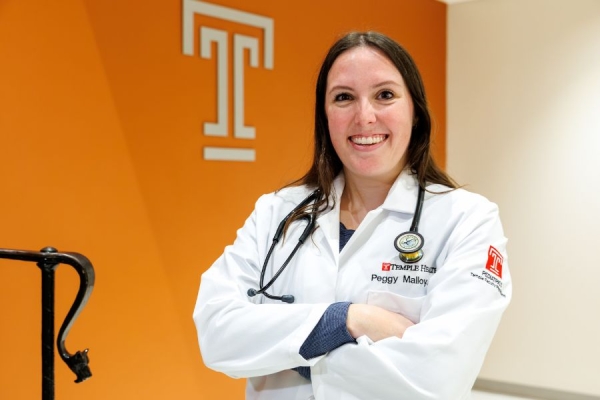 Physcian Assistant Peggy Malloy smiling in her white coat with a stethoscope around her neck stands in front of an orange wall with a Temple logo.