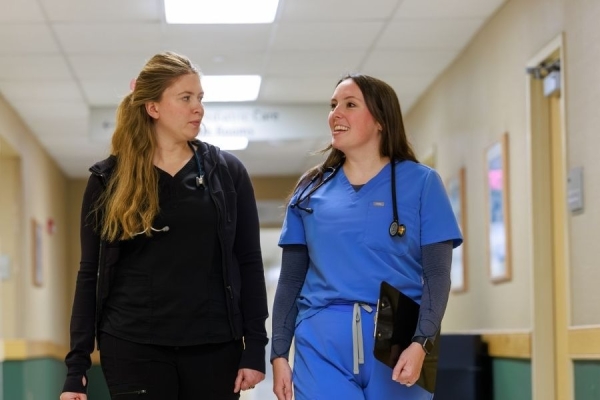 Physician Assistant Peggy Malloy in blue scrubs, holding a clipboard, chats with coworker in black scrubs in a brightly lit hallway lined with green and beige walls, framed posters, and a bulletin board.